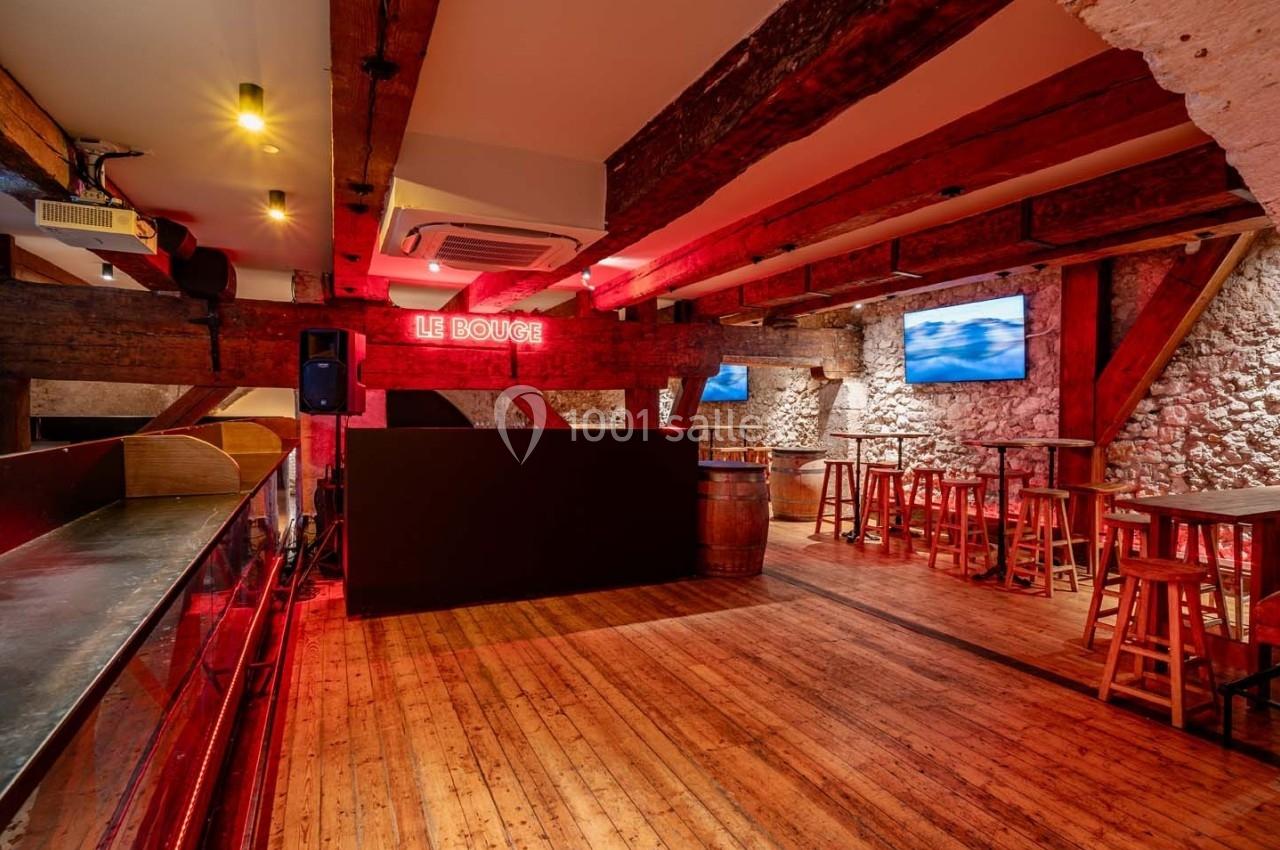 Salle en bois avec poutres apparentes, bar à gauche, tables hautes et écrans accrochés aux murs en pierre.