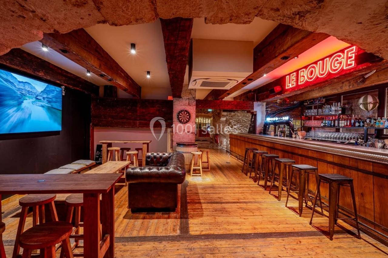 Intérieur d'un bar avec comptoir en bois, tabourets alignés, tables hautes, canapé en cuir et enseigne lumineuse.