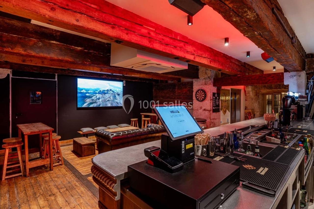 Intérieur d'un bar avec comptoir, tabourets, canapé en cuir, écran mural et éclairage chaleureux.