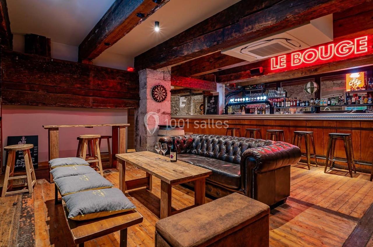Intérieur d'un bar avec canapé en cuir, tables en bois, tabourets et enseigne lumineuse rouge au mur.