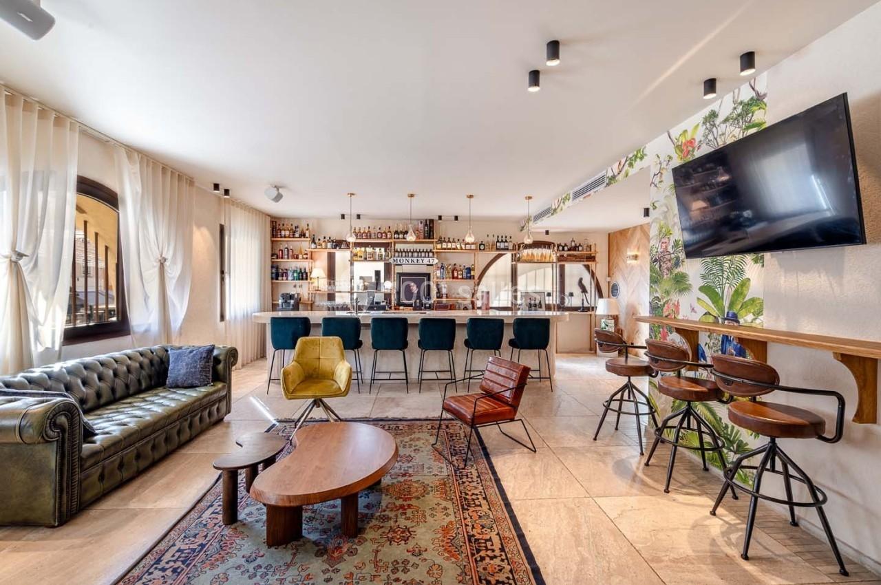 Bar lumineux avec comptoir moderne, étagères de bouteilles, canapés, fauteuils et décoration murale végétale.