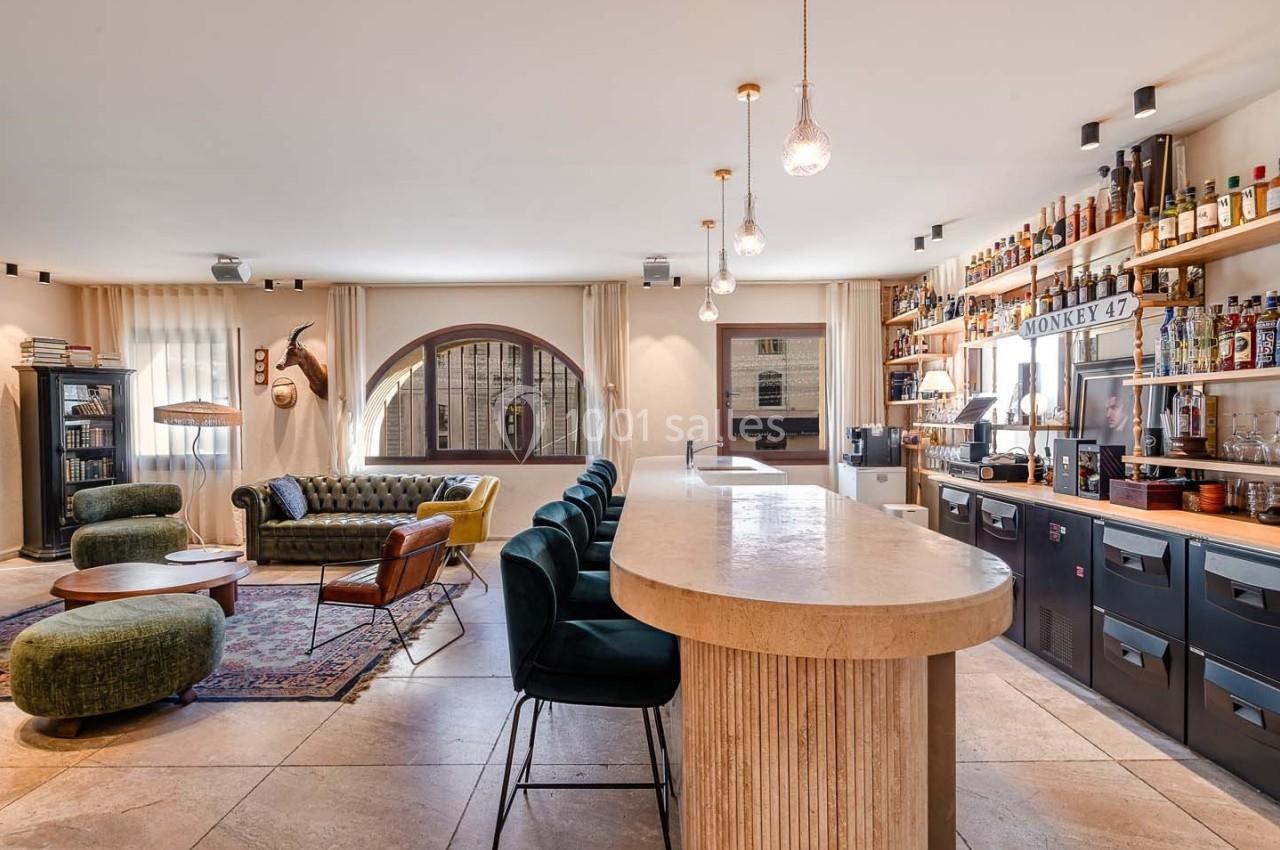 Espace intérieur lumineux avec un bar en bois, des étagères de bouteilles, un salon et de grandes fenêtres arrondies.