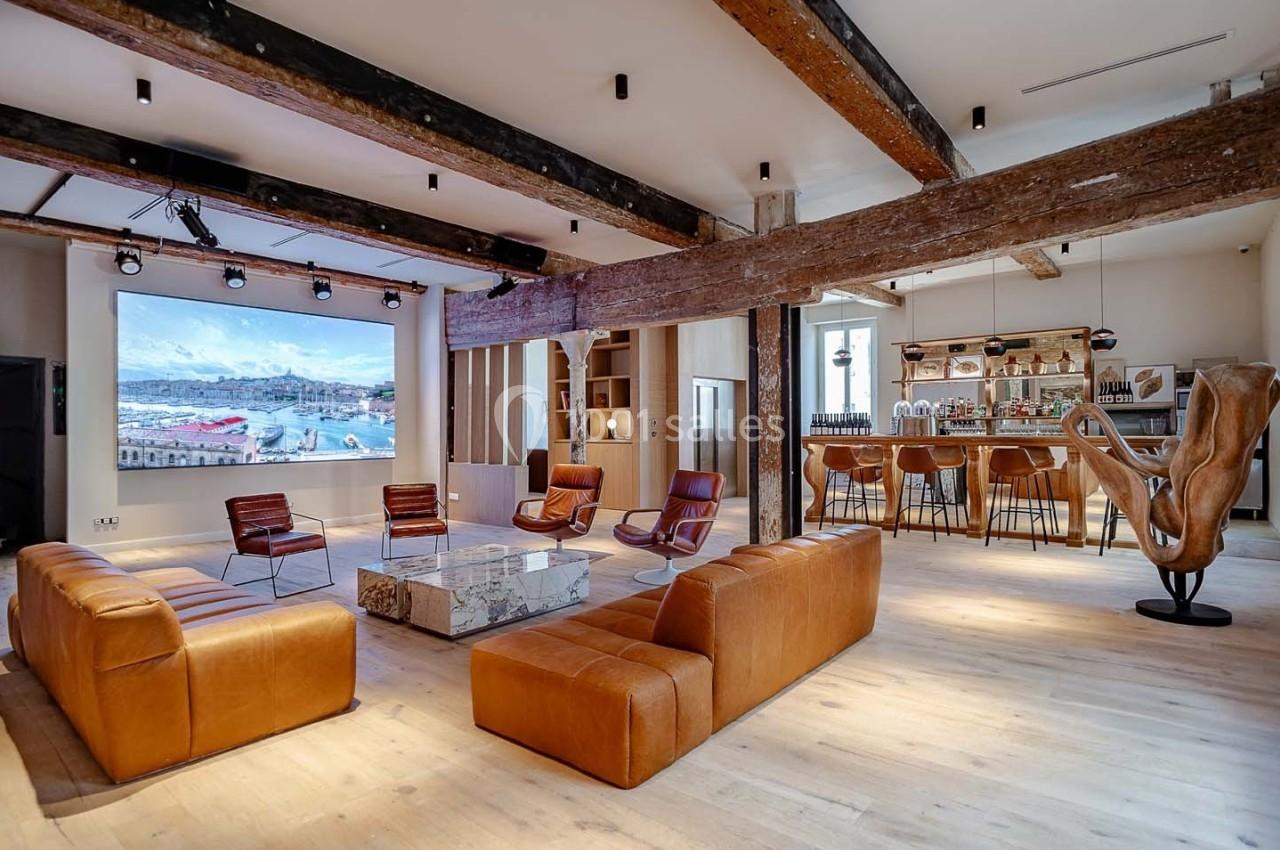 Salon spacieux avec canapés en cuir, table basse en marbre, bar en arrière-plan et poutres apparentes au plafond.