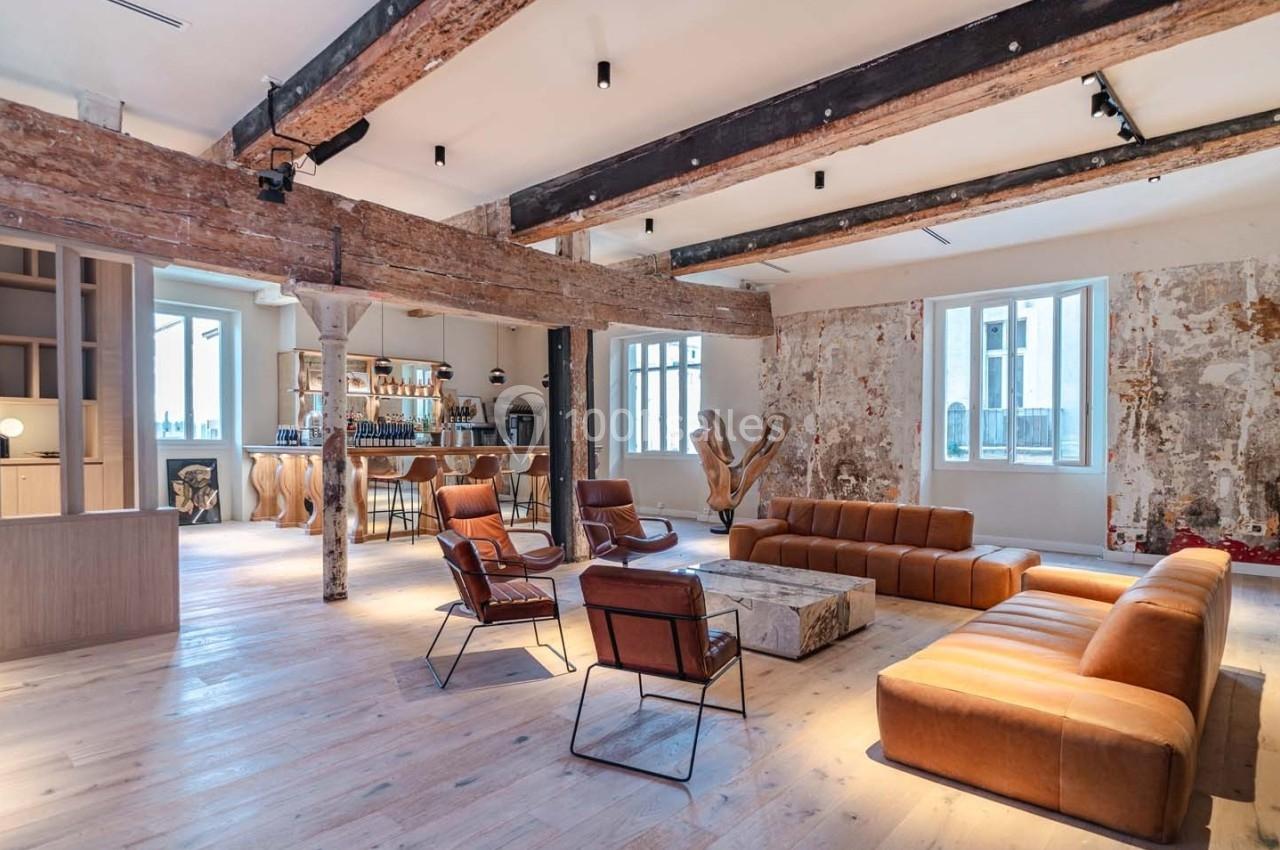 Espace intérieur lumineux avec poutres apparentes, murs texturés, mobilier en cuir et coin bar en arrière-plan.