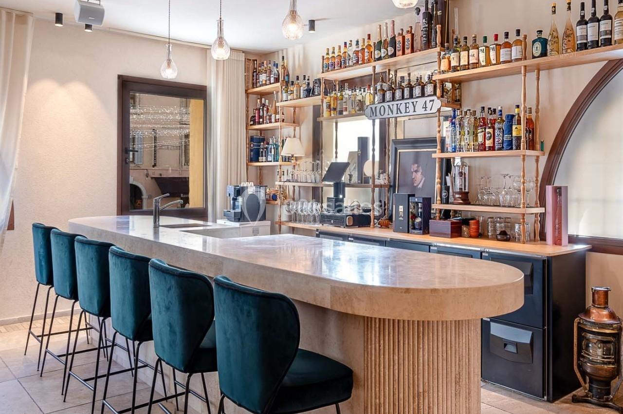 Bar moderne avec comptoir en marbre, tabourets verts, étagères remplies de bouteilles et éclairage suspendu.