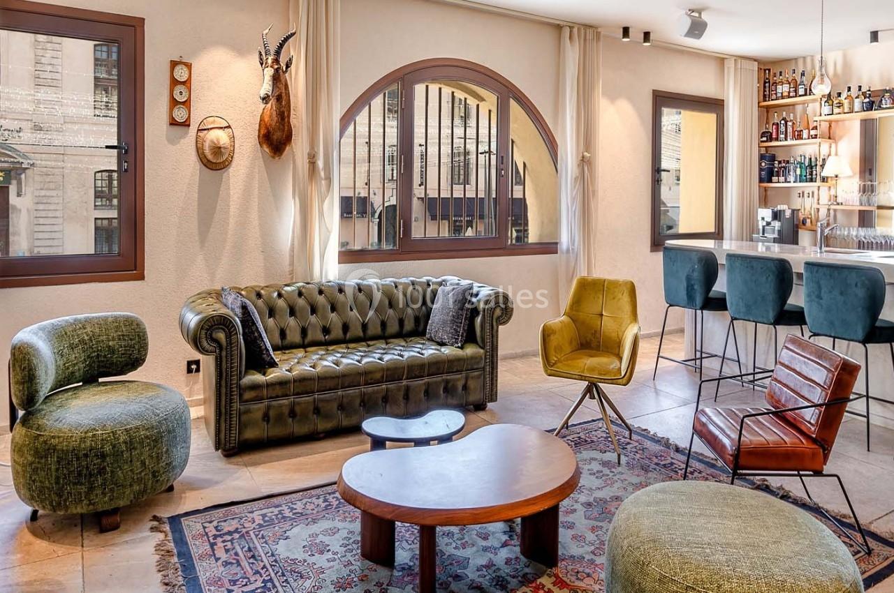 Salon lumineux avec mobilier éclectique, canapé en cuir, fauteuils design, tapis coloré et bar en arrière-plan.