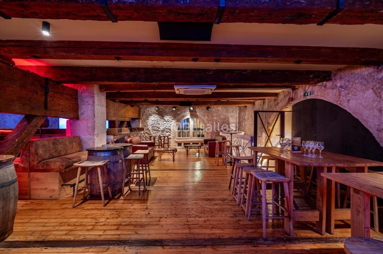 Salle en pierre avec poutres apparentes, mobilier en bois, éclairage chaleureux et grandes fenêtres en arrière-plan.