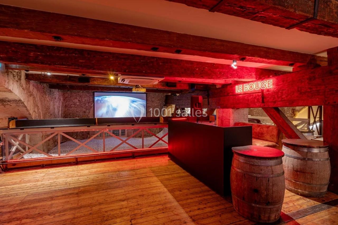 Salle avec poutres en bois apparentes, bar noir, tonneaux en décoration et écran projetant une image lumineuse.