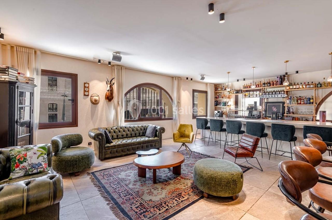 Salon spacieux avec mobilier vintage, bar en arrière-plan, grandes fenêtres et lumière naturelle.