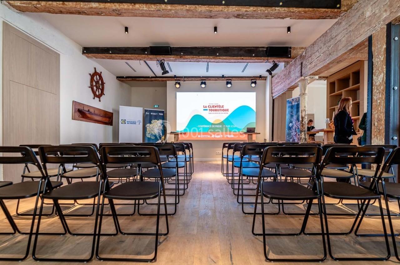 Salle de conférence avec des rangées de chaises noires face à un écran de projection allumé.