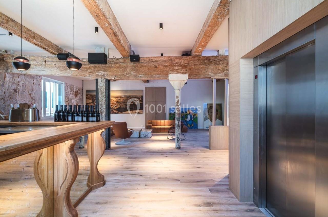 Intérieur lumineux avec poutres apparentes, sol en bois, comptoir sculpté, ascenseur et mobilier moderne.