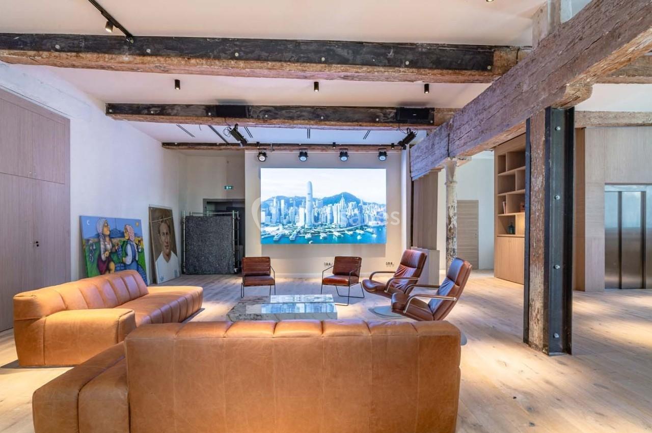 Espace lumineux avec canapés en cuir, écran mural affichant une vue urbaine, poutres apparentes et parquet clair.