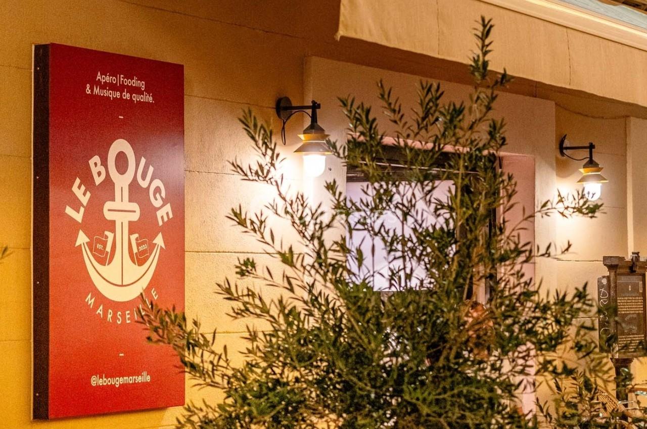 Enseigne rouge d'un établissement avec un logo d'ancre, éclairée par des lampes murales, partiellement masquée par un…