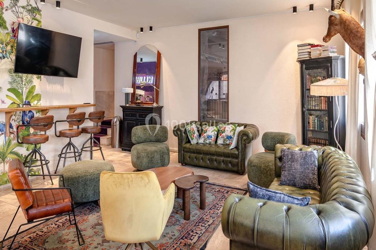 Salon confortable avec canapés en cuir vert, fauteuils colorés, tapis persan, bar avec tabourets et décoration éclectique.