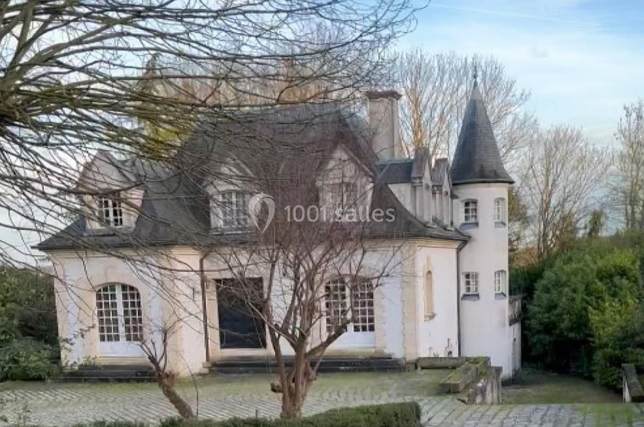 Grande maison blanche avec des toits en ardoise, entourée d'arbres et d'une allée pavée bordée de haies.
