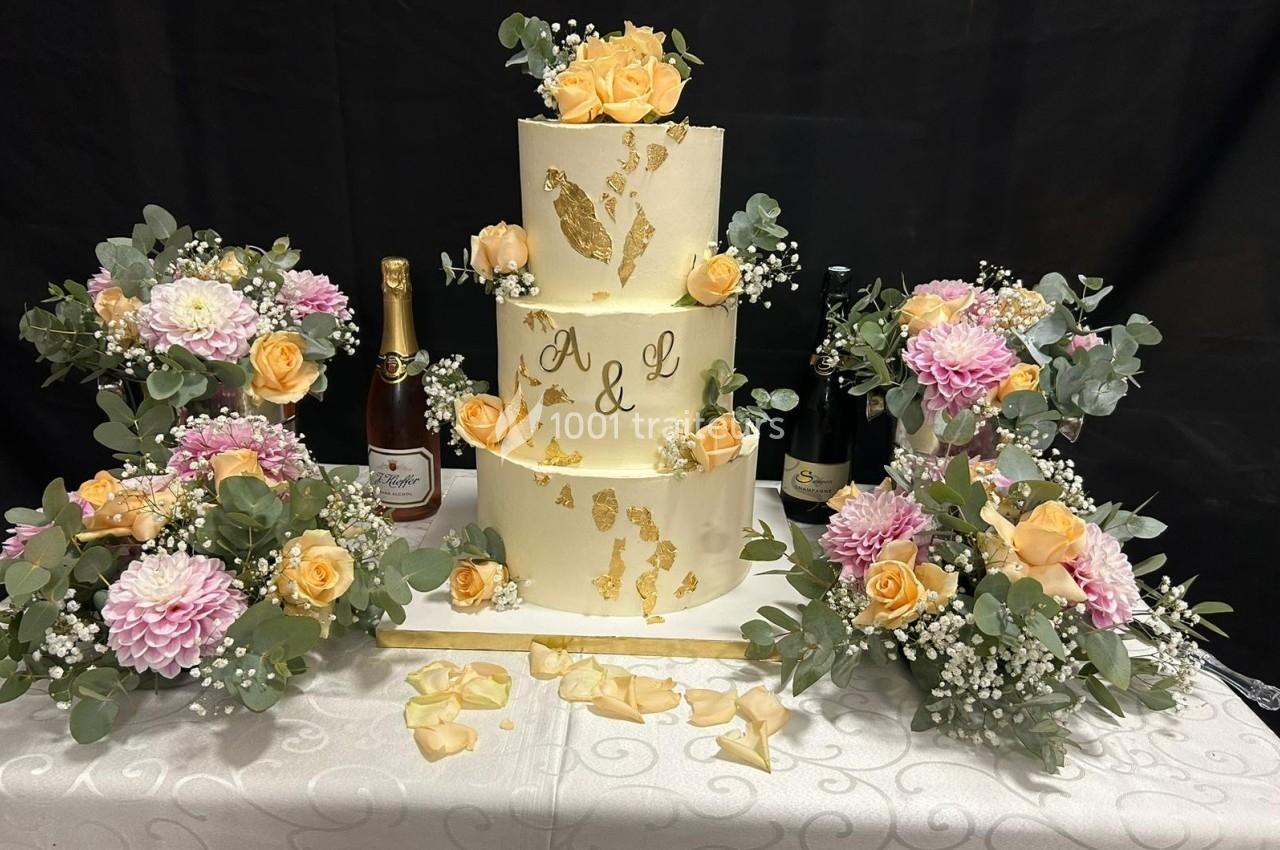 Gâteau de mariage à trois étages décoré de fleurs, entouré de bouteilles de vin et d'arrangements floraux.
