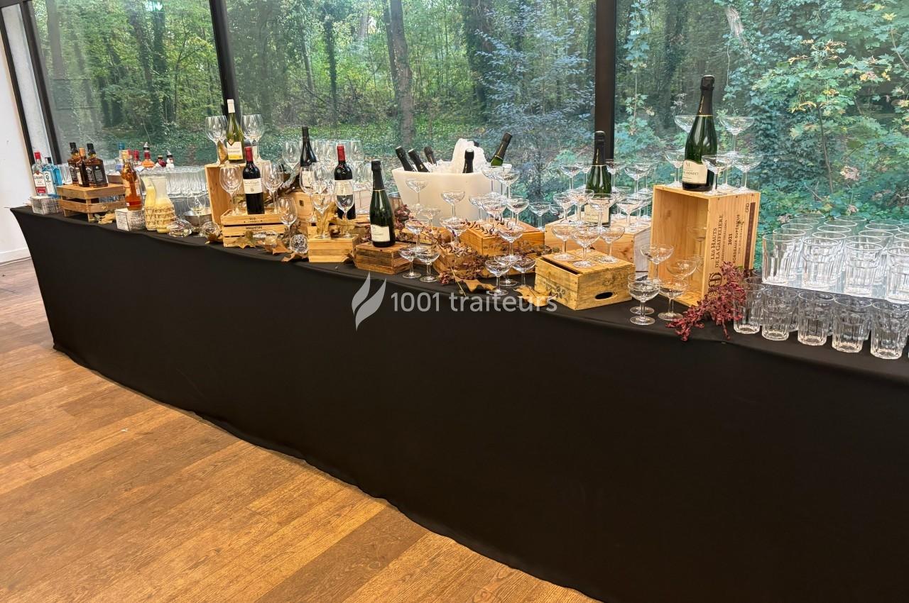 Table de bar avec bouteilles d'alcool, verres et caisses en bois, disposée devant une baie vitrée donnant sur un jardin…