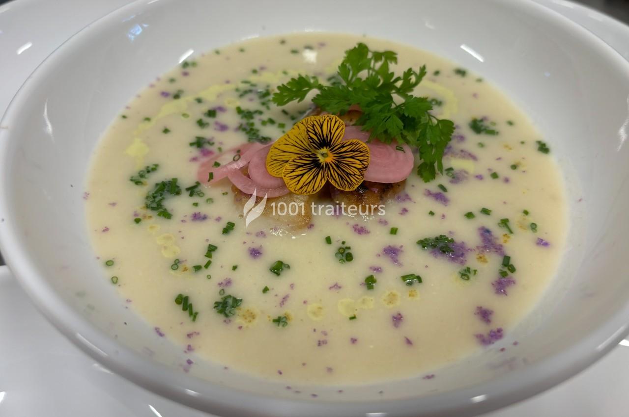 Soupe crémeuse garnie de fleurs comestibles, herbes fraîches, pickles et croûtons dans une assiette blanche.