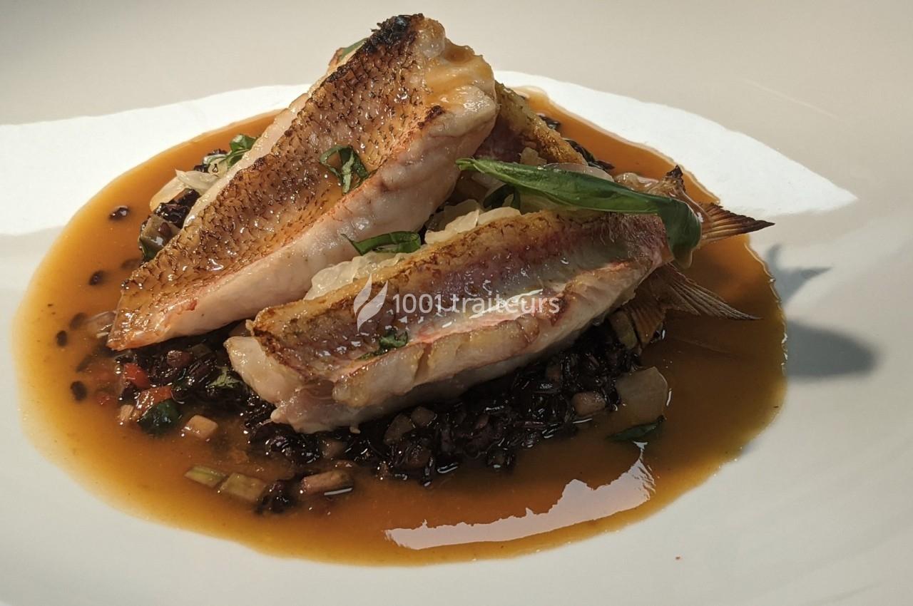 Filets de poisson grillés servis sur un lit de légumes et de riz noir, nappés d'une sauce brune.