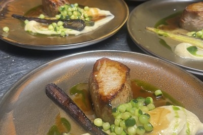 Un chef en tablier marron sourit, bras croisés, dans une cuisine moderne lumineuse.