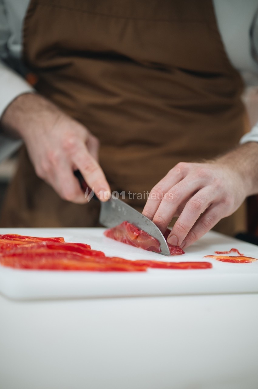 Un cuisinier découpe des poivrons rouges en fines lamelles sur une planche blanche.