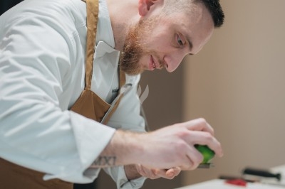 Un chef en tablier marron sourit, bras croisés, dans une cuisine moderne lumineuse.