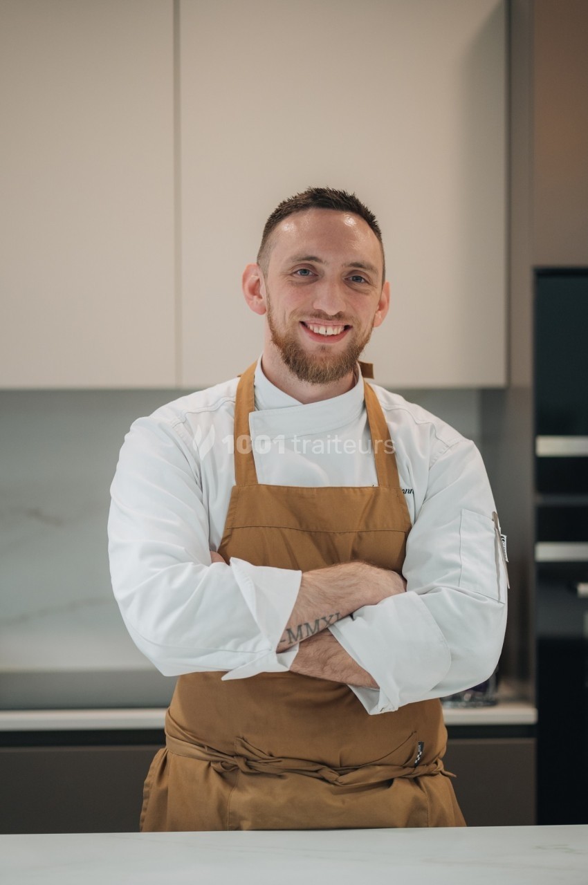 Un chef en tablier marron sourit, bras croisés, dans une cuisine moderne lumineuse.