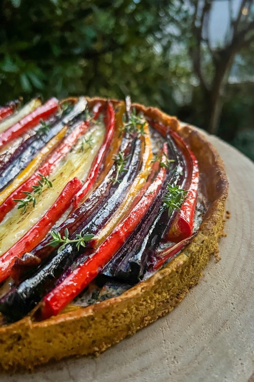 Tarte salée garnie de légumes colorés disposés en rangées sur une pâte dorée, présentée en extérieur.
