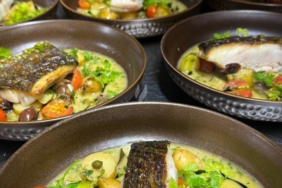 Un chef en tablier marron croise les bras devant un plan de travail dans une cuisine moderne.