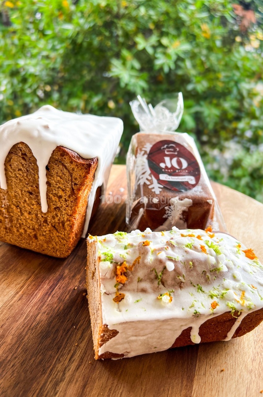 Trois cakes glacés, dont un partiellement tranché, posés sur une planche en bois devant un fond végétal.