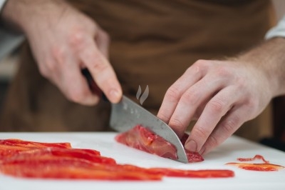 Un chef en tablier marron croise les bras devant un plan de travail dans une cuisine moderne.