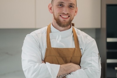 Un chef en tablier marron croise les bras devant un plan de travail dans une cuisine moderne.
