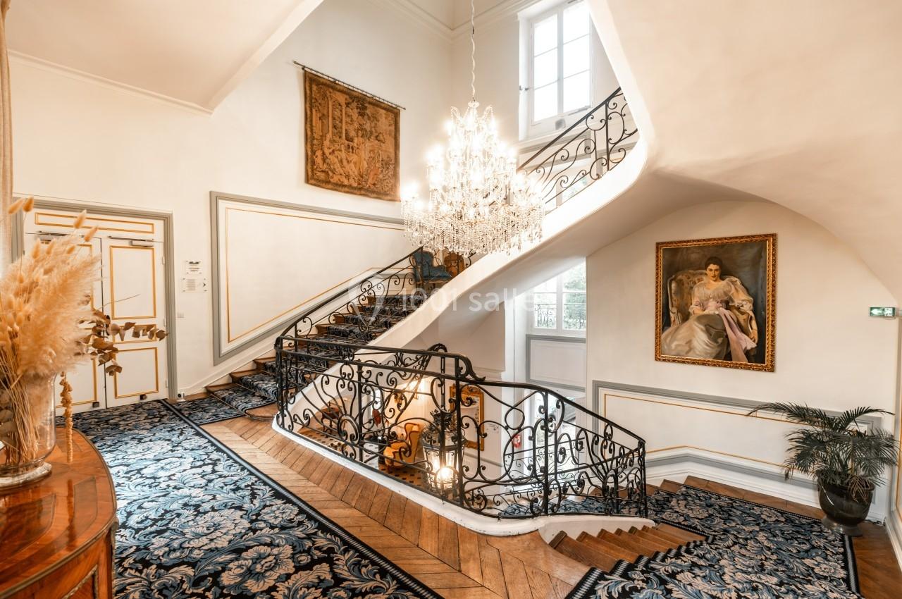 Escalier central élégant avec rampe en fer forgé, tapis décoratif, lustre imposant et tableaux accrochés aux murs.