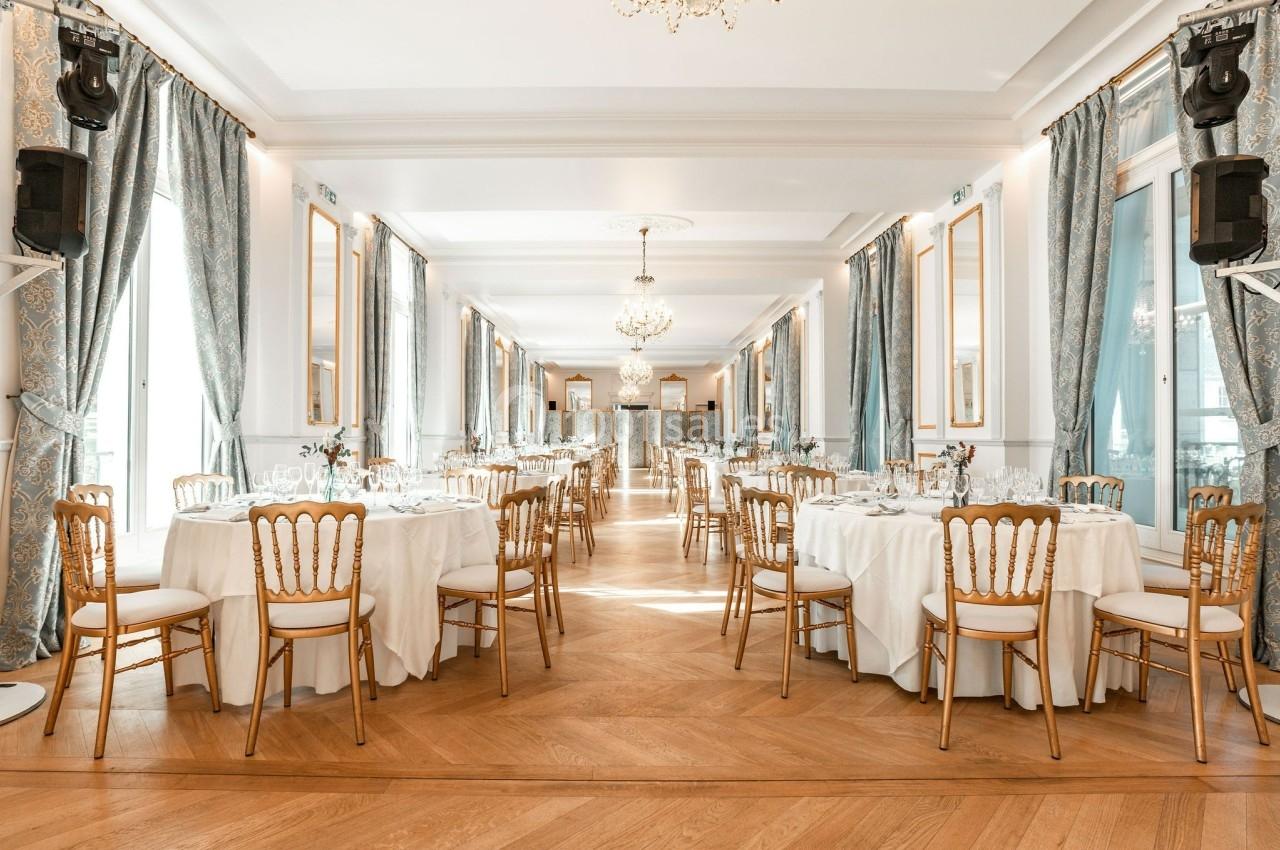 Salle de réception élégante avec tables rondes dressées, chaises dorées et grandes fenêtres encadrées de rideaux bleus.