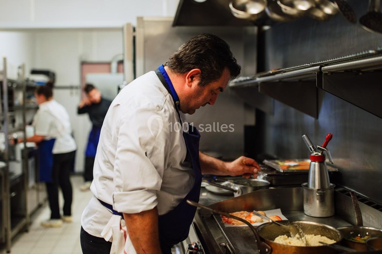 Un chef en tablier bleu prépare un plat dans une cuisine professionnelle, avec d'autres cuisiniers en arrière-plan.