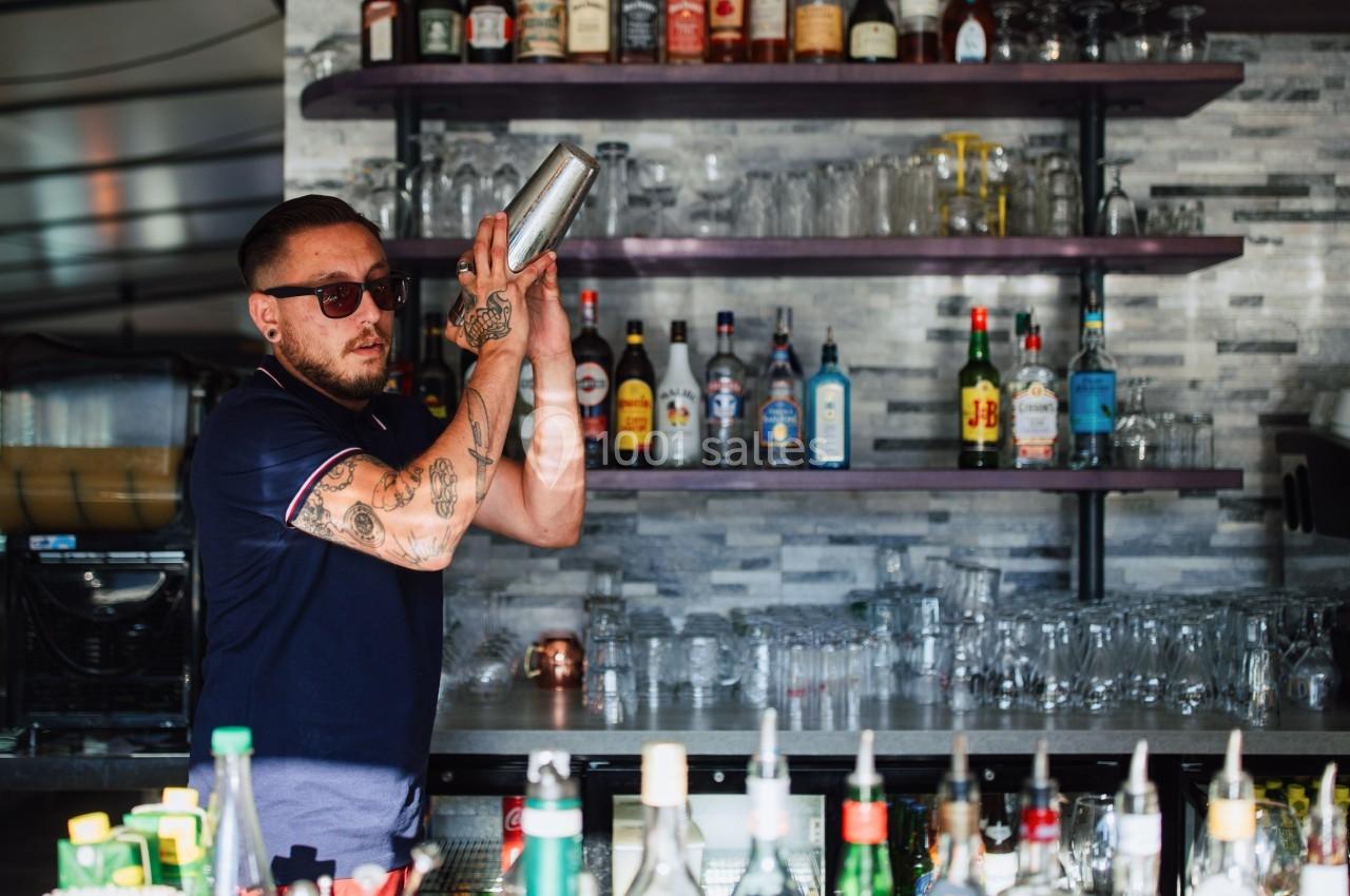 Un barman tatoué mélange un cocktail avec un shaker derrière un comptoir, devant des étagères remplies de bouteilles et de…