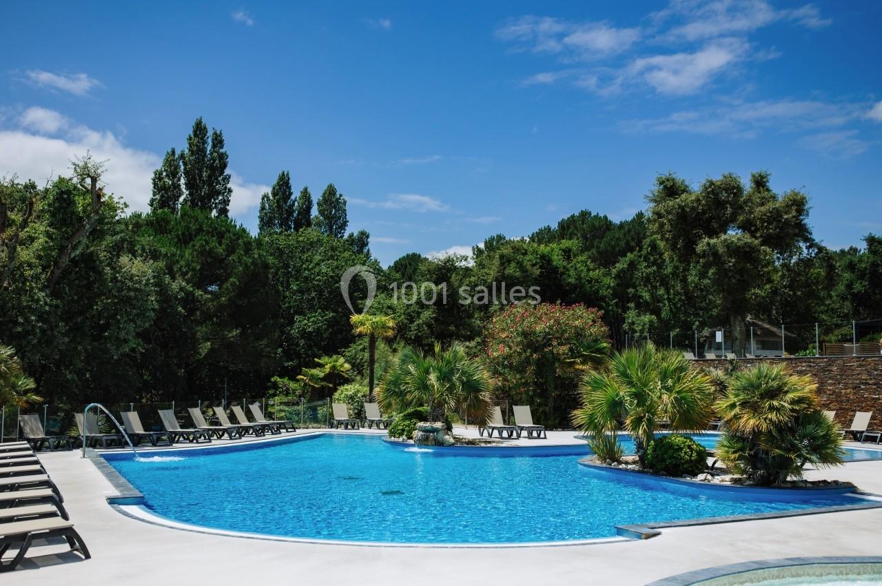 Piscine extérieure entourée de transats, palmiers et végétation, sous un ciel bleu dégagé.