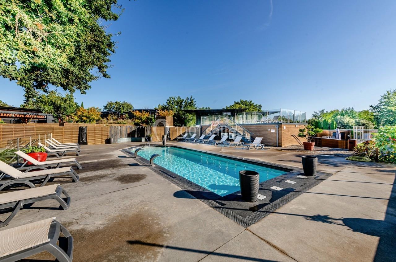 Piscine extérieure entourée de chaises longues, avec des arbres et un ciel dégagé en arrière-plan.