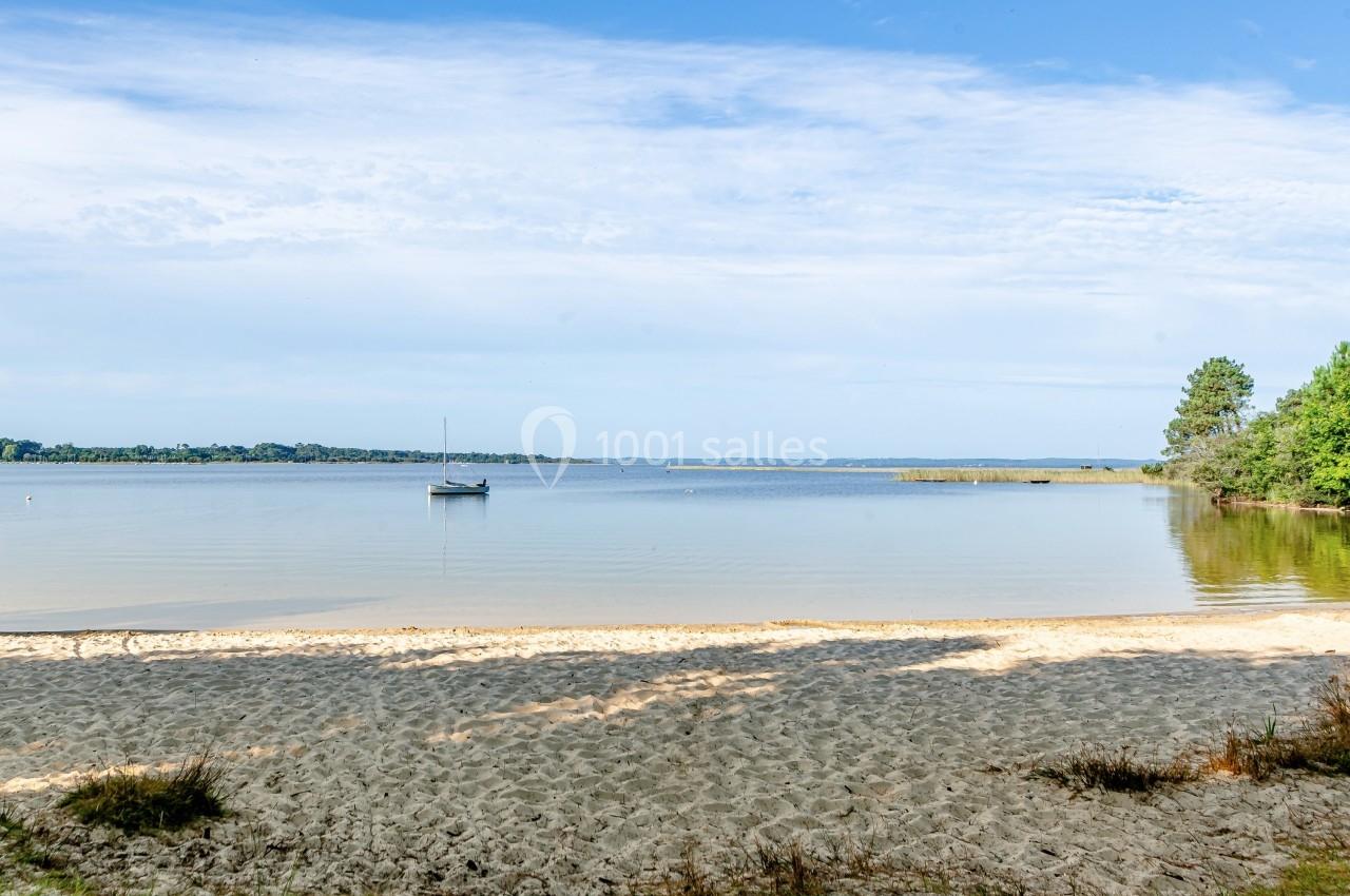 Plage de sable bordant un lac calme avec un voilier ancré au loin et des arbres en arrière-plan.