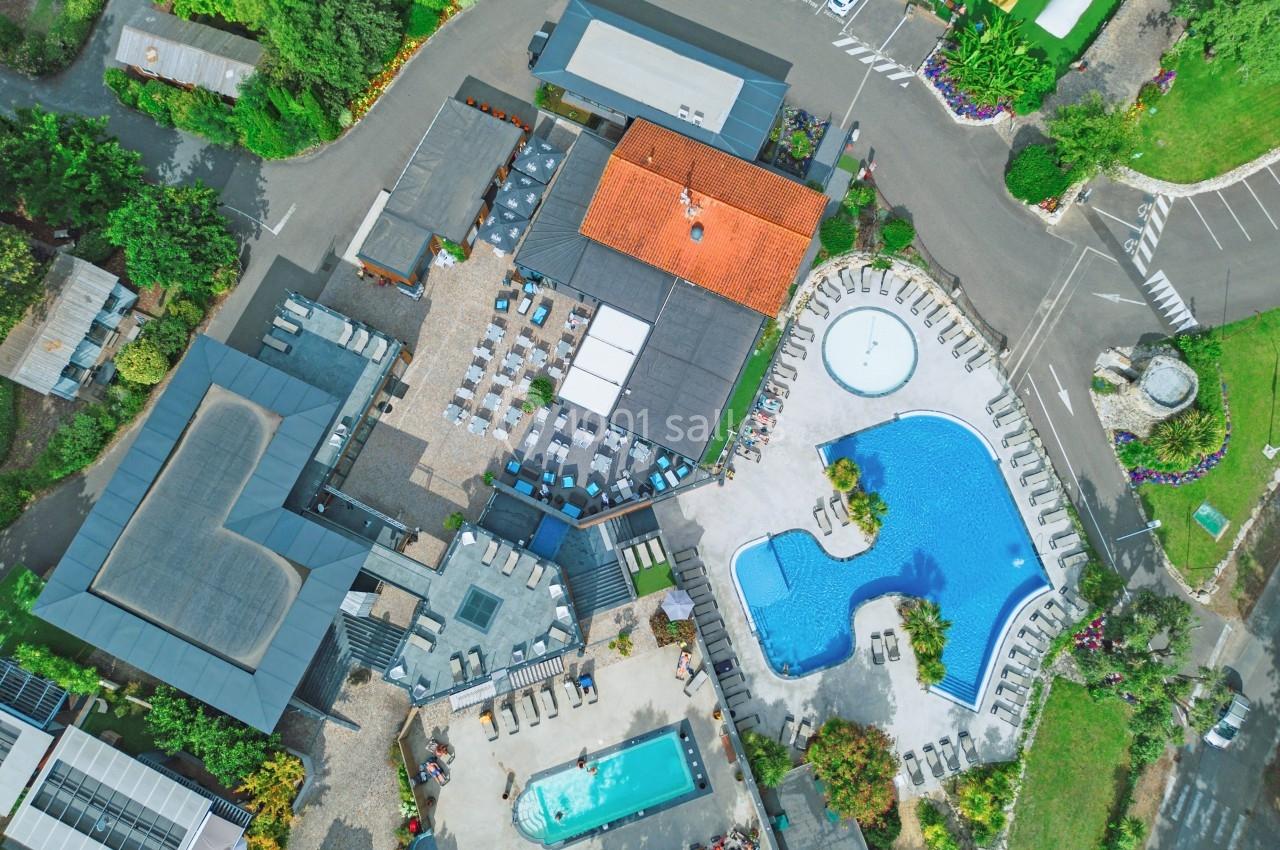 Vue aérienne d'un complexe avec piscines, bâtiments, espaces verts et parkings, entouré de végétation.