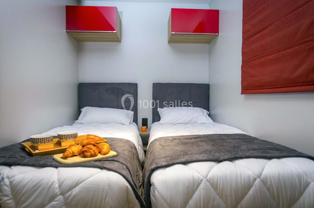 Deux lits simples avec couvertures grises, un plateau de petit-déjeuner posé sur l'un, étagères rouges au mur.