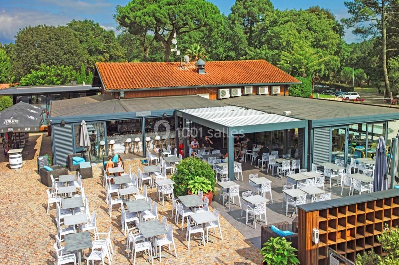 Terrasse d'un restaurant avec tables et chaises en extérieur, entourée de végétation et d'arbres sous un ciel dégagé.
