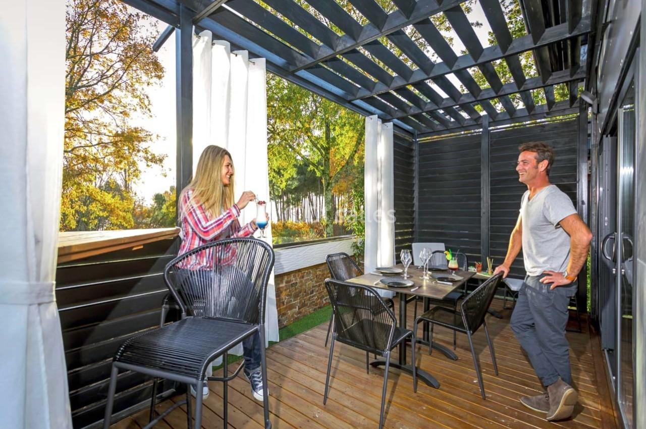 Une femme et un homme discutent sur une terrasse en bois couverte, aménagée avec une table et des chaises.