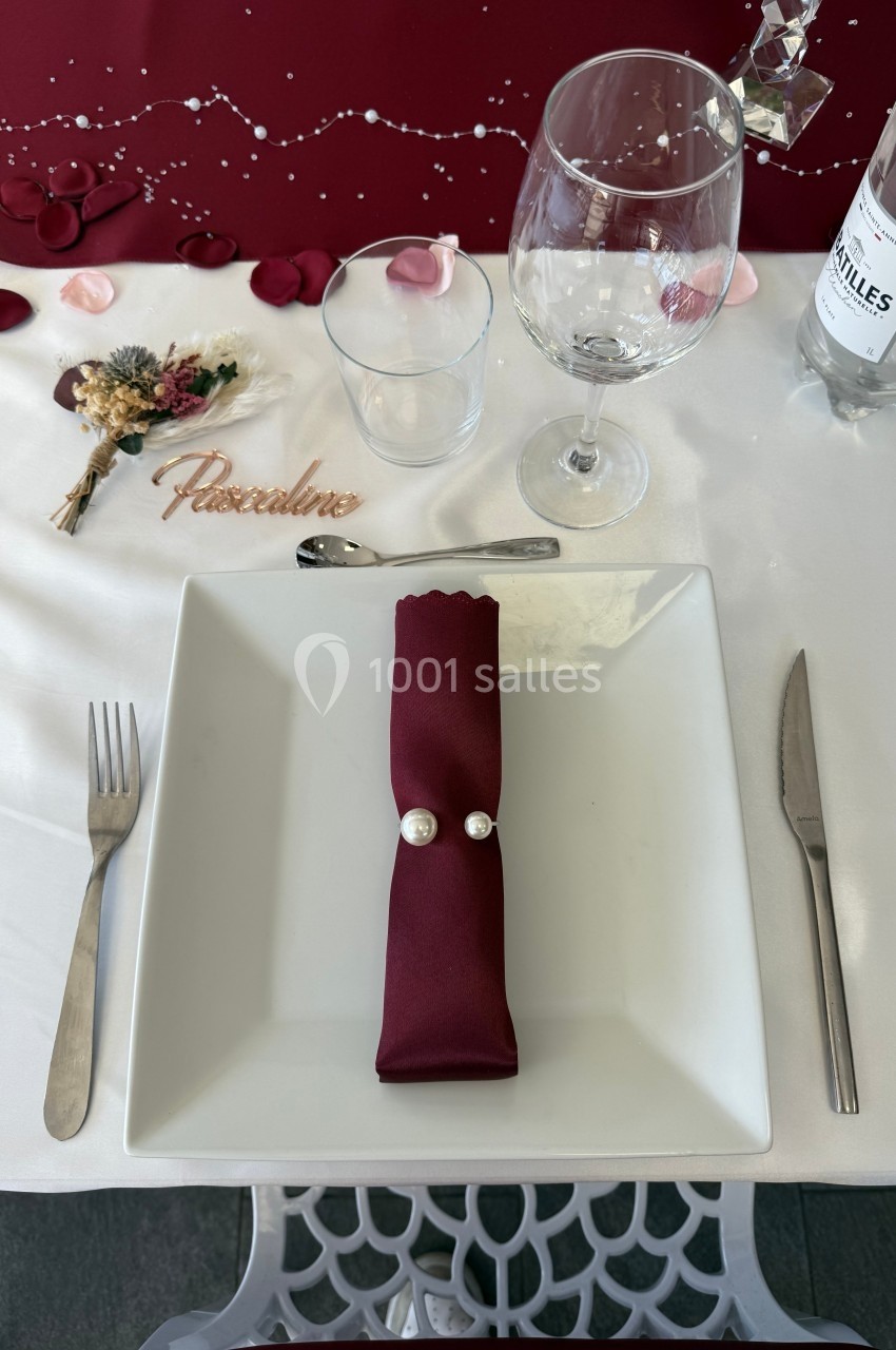 Serviette rouge pliée avec deux perles posée sur une assiette blanche, entourée de couverts et d'un verre à pied.