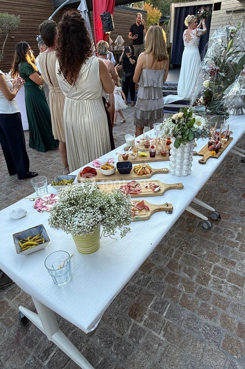 Table garnie de planches apéritives et décorations florales lors d'un événement en plein air avec des invités debout.