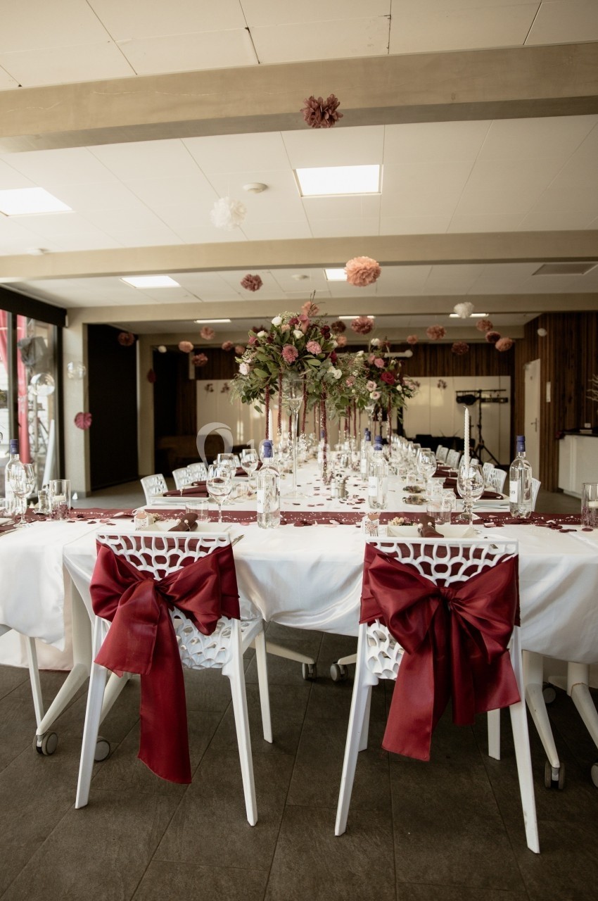 Table décorée pour un événement avec nappes blanches, chaises ornées de nœuds rouges et centre de table floral.