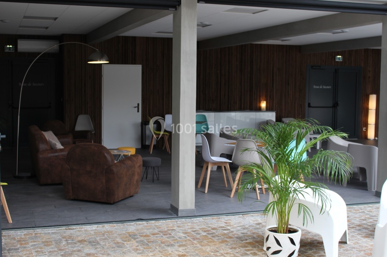Espace intérieur moderne avec fauteuils, chaises, tables et plantes, éclairé par une lumière naturelle.