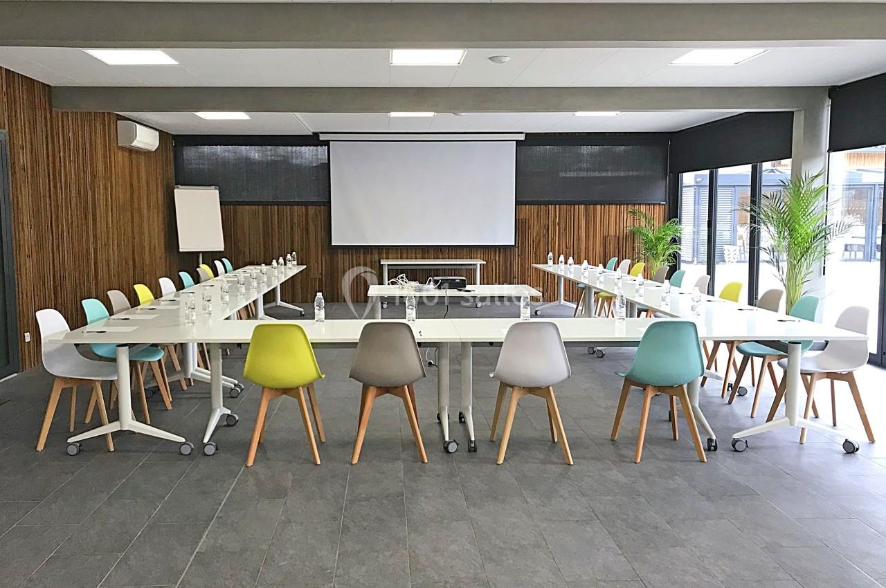 Salle de réunion lumineuse avec tables en U, chaises colorées, écran de projection et plantes en arrière-plan.