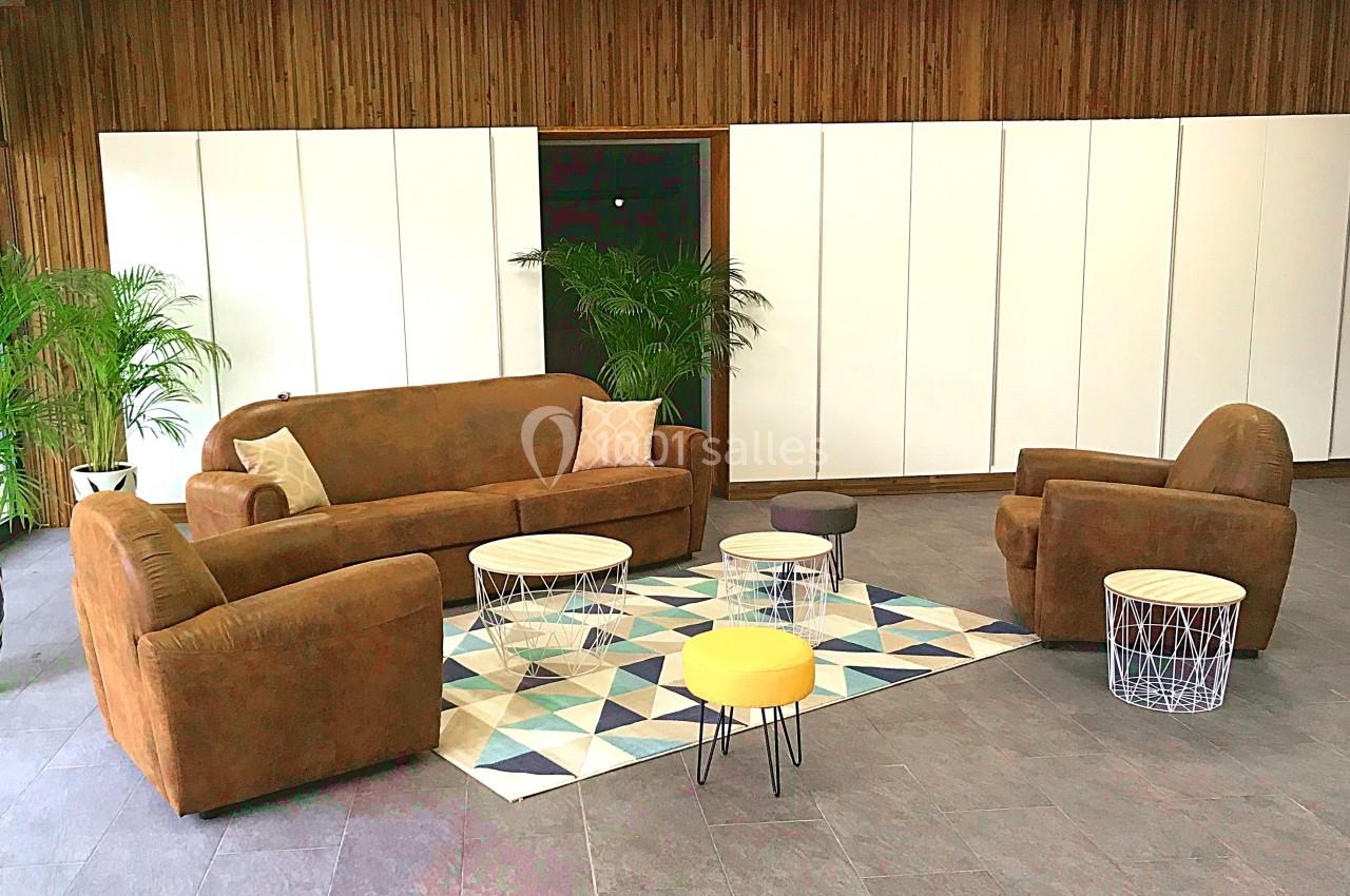 Salon avec canapé, fauteuils en velours marron, tables basses modernes et tapis géométrique, entouré de plantes vertes.