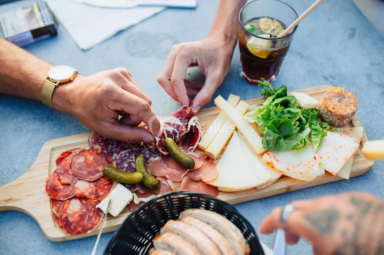 Planche de charcuterie et fromages variés avec cornichons, salade et pain, servie sur une table avec boissons.